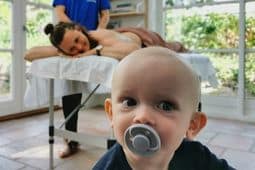 Massør giver efterfødselsmassage til en klient og barnet kigger ind i kameraet